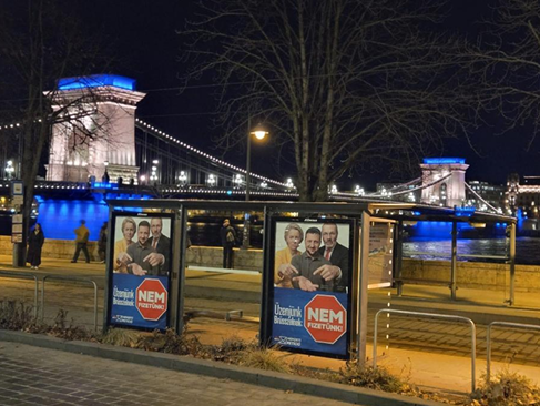 Фото трамвайної зупинки з плакатами, зроблене 24 лютого 2026 року на тлі Ланцюгового мосту в Будапешті, підсвіченого синьо-жовтими кольорами на знак солідарності з Україною (жест мерії Будапешта). Типовий слоган: «Послання Брюсселю — ми НЕ будемо платити!». Центр стратегічних комунікацій SPRAVDI