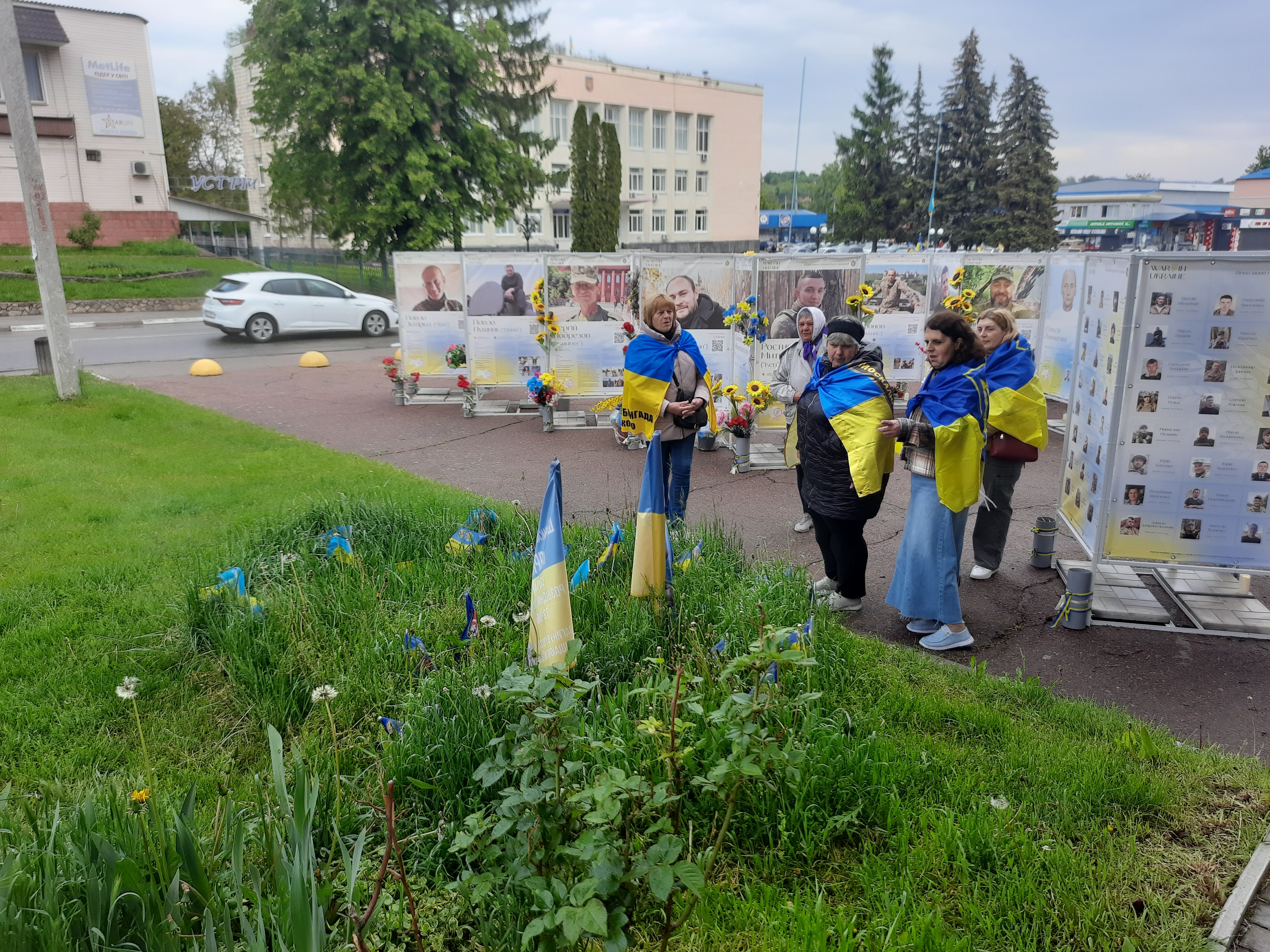 Матері полеглих воїнів стоять біля прапорців, поставлених на міському газоні. Комунальники цю ділянку оминають увагою і не доглядають. Фото авторки